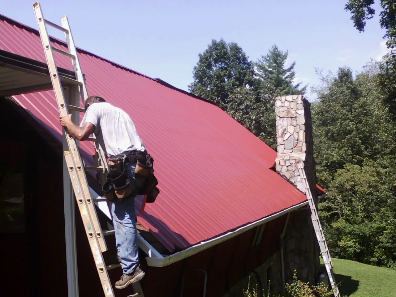 Roofer on a ladder beside a red metal roof and stone chimney — Soffit & Fascia Repair by Roofing Brothers in Roslyn